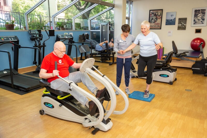 Senior man using a recumbent bike and senior woman working on balance exercises with personal trainer in the fitness center