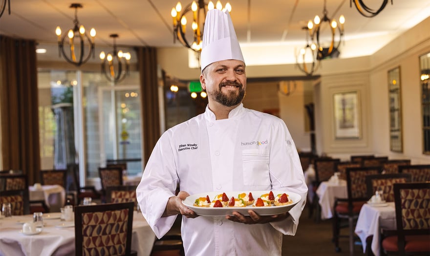 Chef in white coat and hat holding plate of food in the dining room at Piedmont Gardens