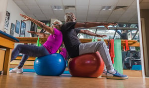 Senior man and woman sitting back-to-back on fitness balls in the Piedmont Gardens fitness center