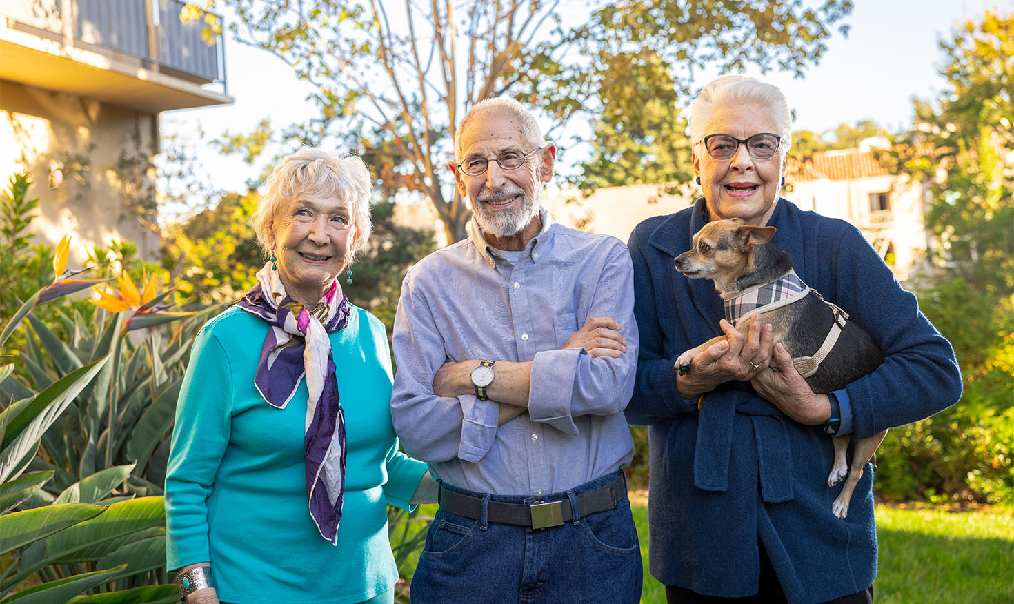 Two senior women — one holding a small dog — and a senior man standing outside