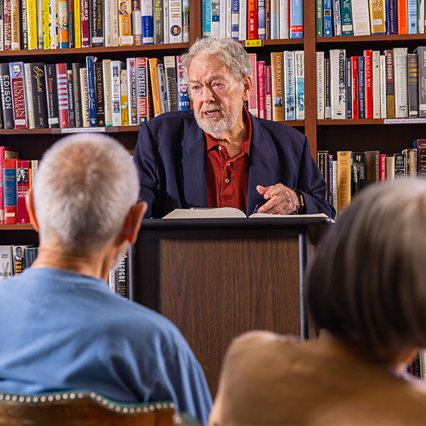 Senior man at a podium in front of bookshelves lecturing to crowd