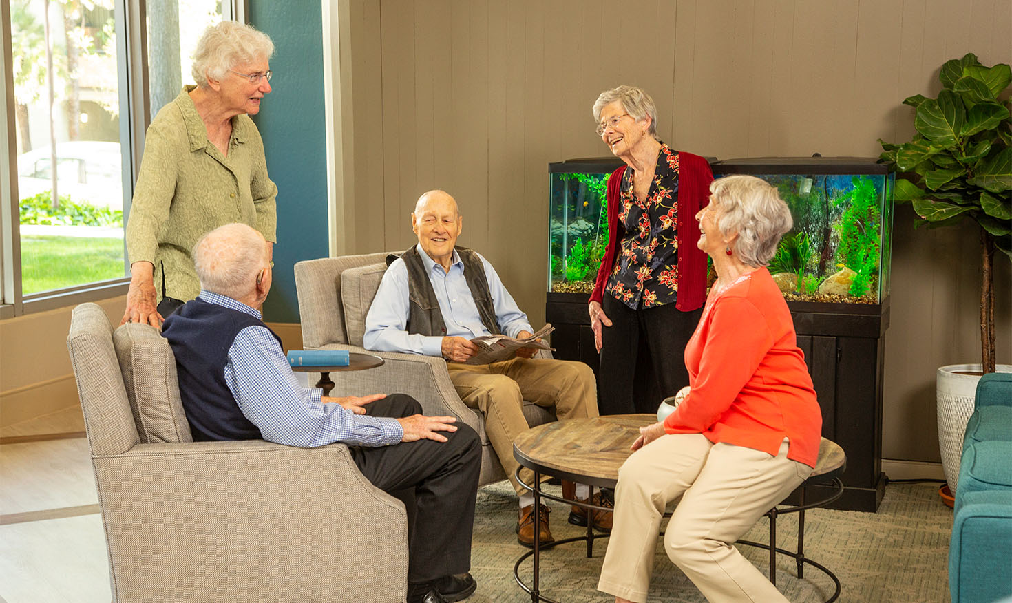 Three senior women and two senior men standing and sitting in a common area with a fish tank at Piedmont Gardens