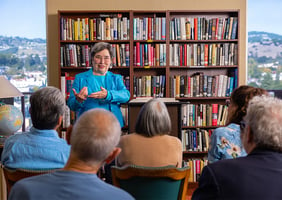 Senior woman at a podium in front of bookshelves lecturing to crowd of seniors