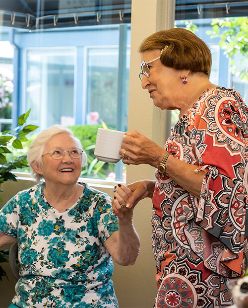 Two senior women, one sitting and one standing, chatting and holding coffee while having coffee