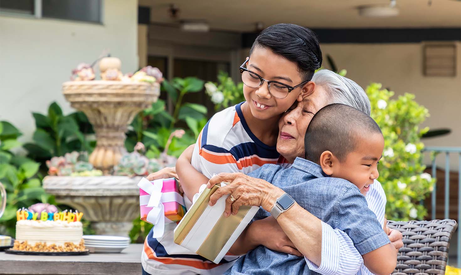 Senior woman holding wrapped gifts and hugging her two grandsons at an outdoor birthday party