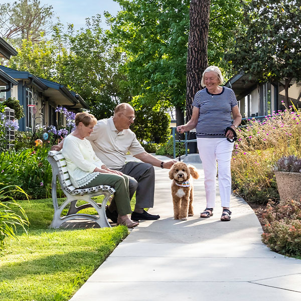 Senior woman walking dog past a senior couple sitting on an outdoor bench at Redwood Terrace