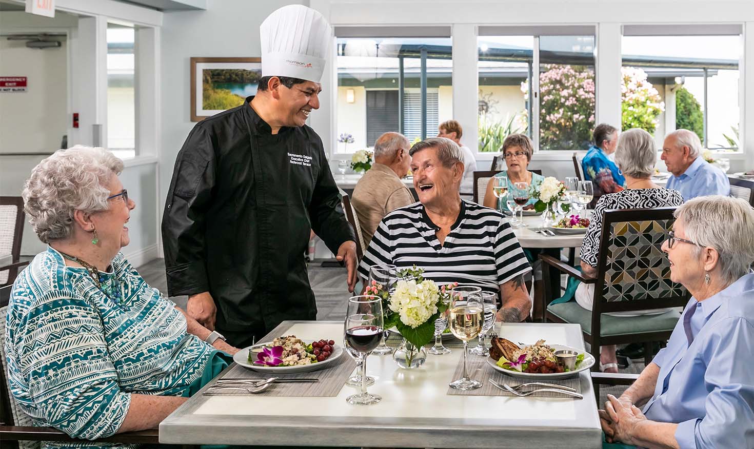 Group of seniors sitting down for a meal and chatting with the chef in the dining room at Redwood Terrace