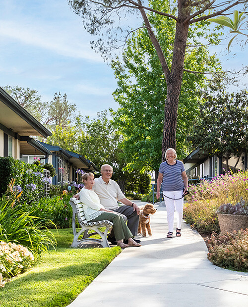 Senior woman walking dog past a senior couple sitting on an outdoor bench at Redwood Terrace