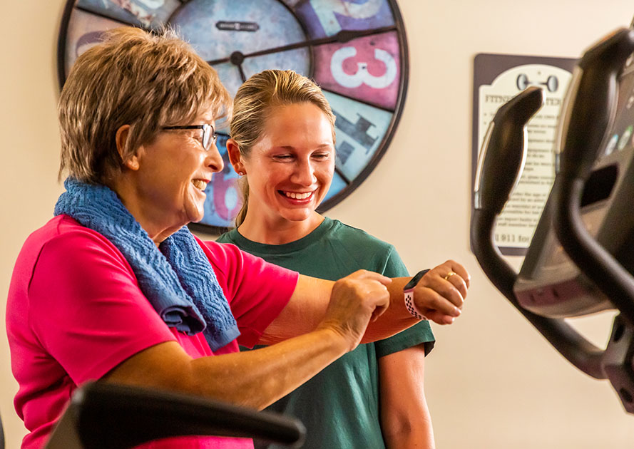 Senior woman looking at her smartwatch and standing next to exercise equipment with a personal trainer 