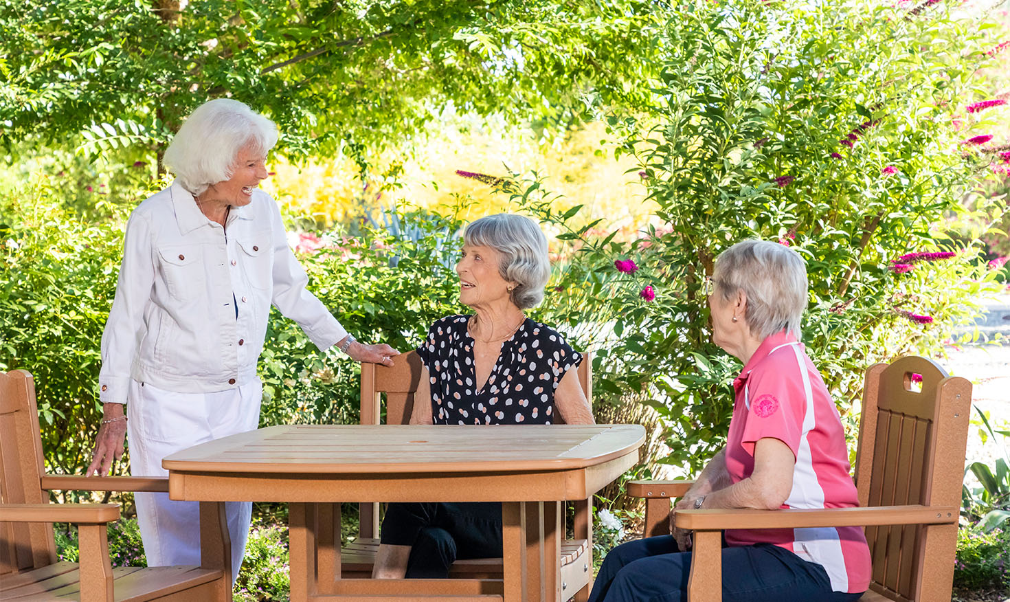 Senior woman standing and talking to two senior women sitting at an outdoor table in the garden at Rosewood