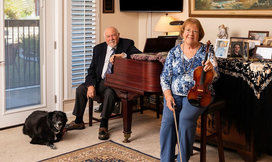 Senior man sitting at a piano and senior woman holding a violin with their dog on the floor in their apartment home at Rosewood