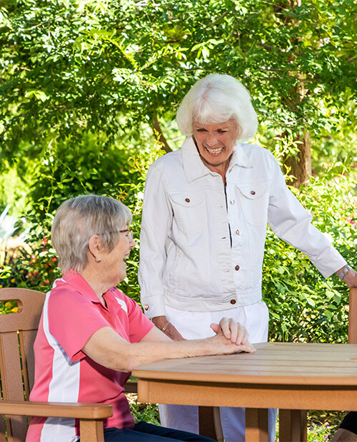 Senior woman standing and talking to another senior women sitting at an outdoor table in the garden at Rosewood