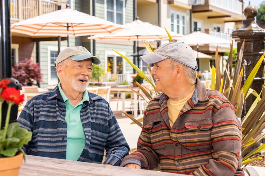 Two senior men wearing baseball caps sitting at an outdoor table at The Terraces at Los Altos