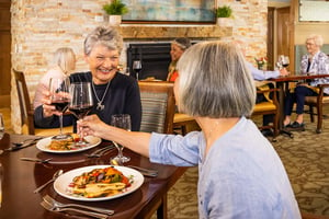 Two senior women clinking glasses of red wine and eating dinner in the fine dining restaurant at The Terraces at Los Altos