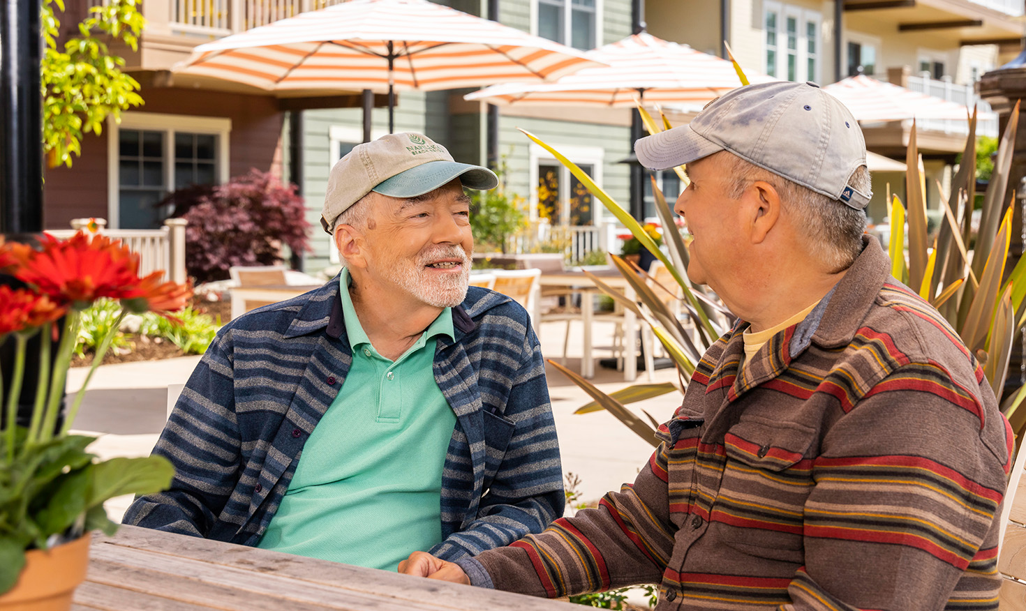 Two senior men wearing baseball caps sitting at an outdoor table at The Terraces at Los Altos