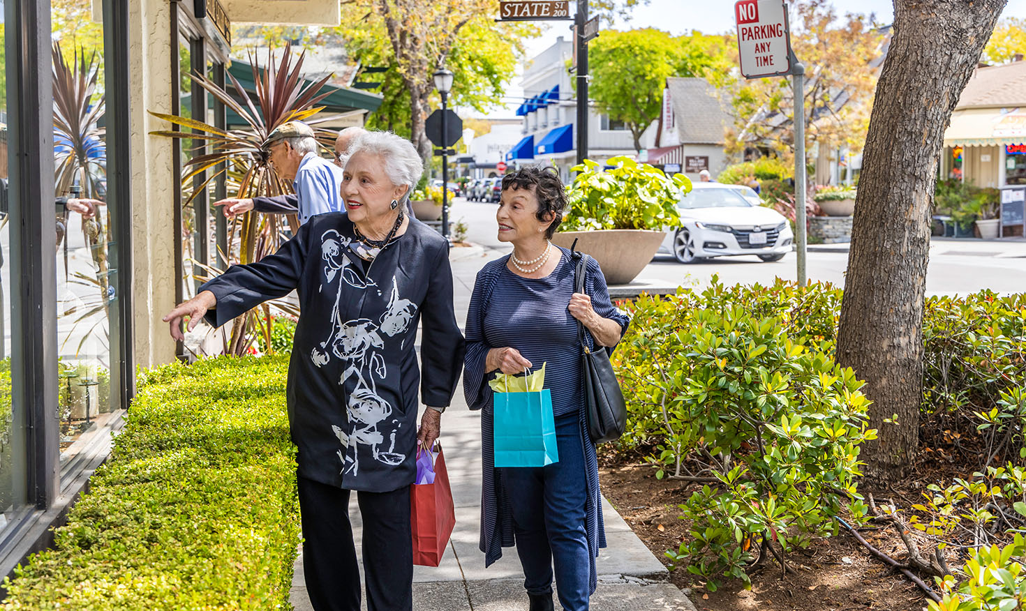 Two senior women carrying shopping bags and walking downtown while one points in a store window