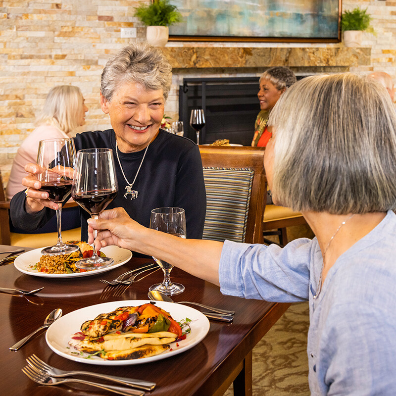 Two senior women clinking glasses of red wine and eating dinner in the fine dining restaurant at The Terraces at Los Altos