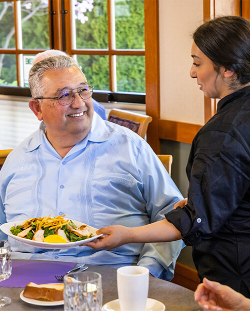 Dining room employee presenting plate of food to a senior man in the dining room at The Terraces at Summitview