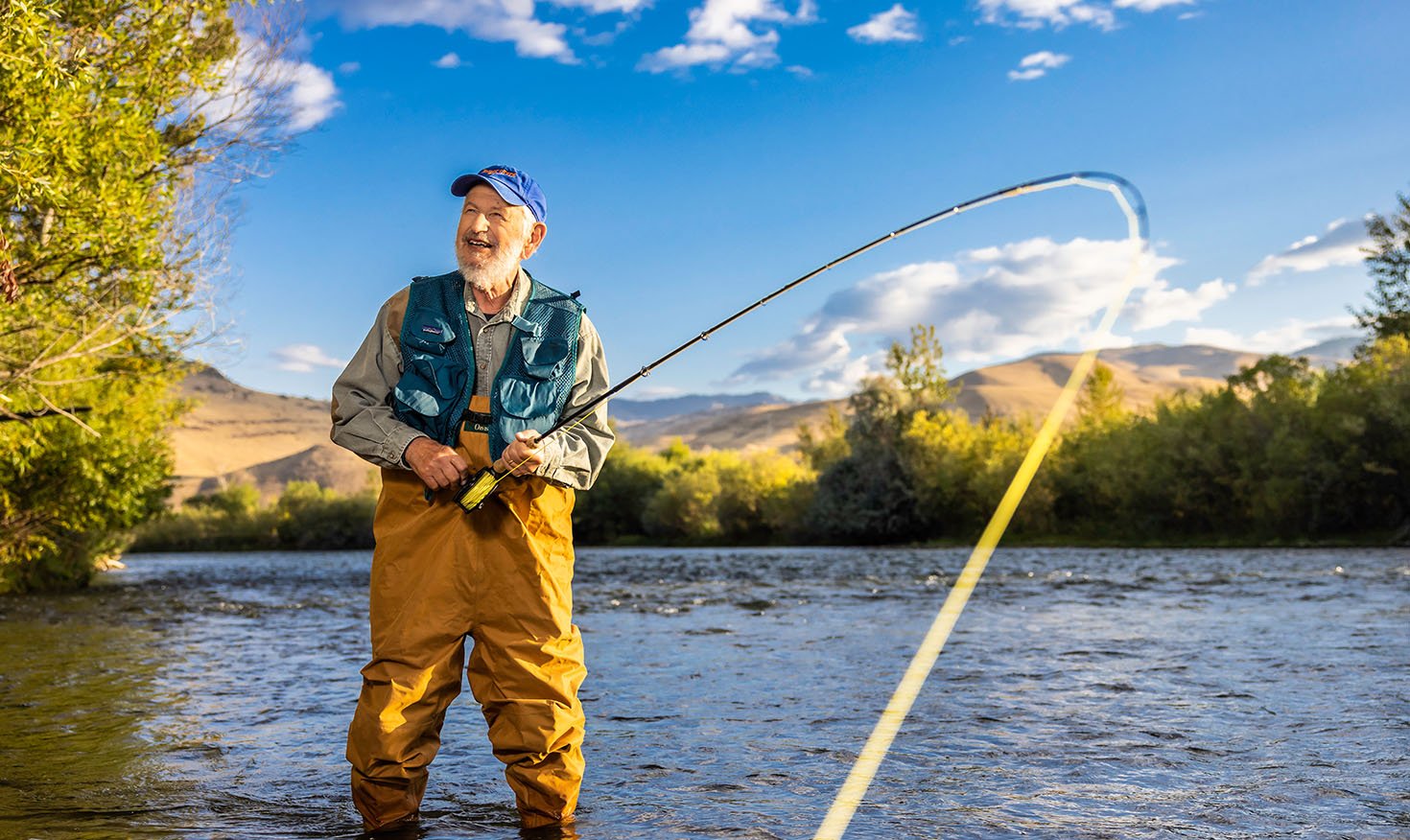 Senior man in waders, a fishing vest and baseball cap standing in a lake fishing