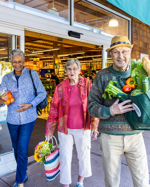 Three seniors leaving a grocery store carrying bags of produce
