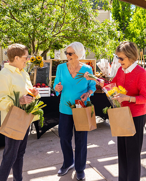 Three senior women shopping at a farmers market carrying paper bags of produce and flowers
