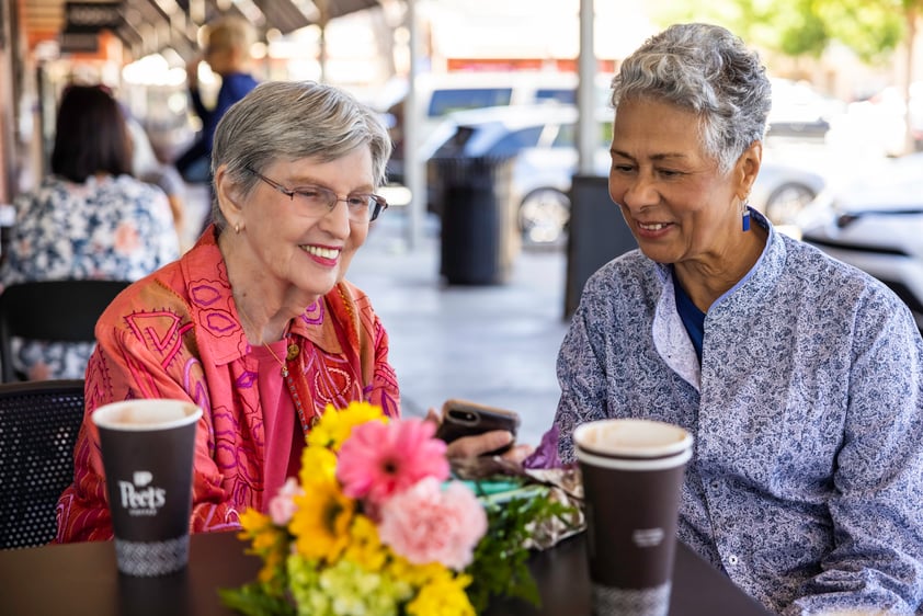 Two senior women enjoying coffee and looking at a cellphone on a patio