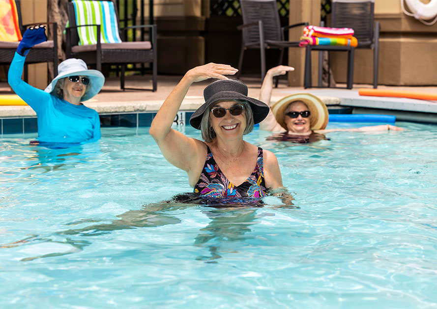 Three senior women wearing sunglasses and sun hats doing water aerobics in an outdoor pool