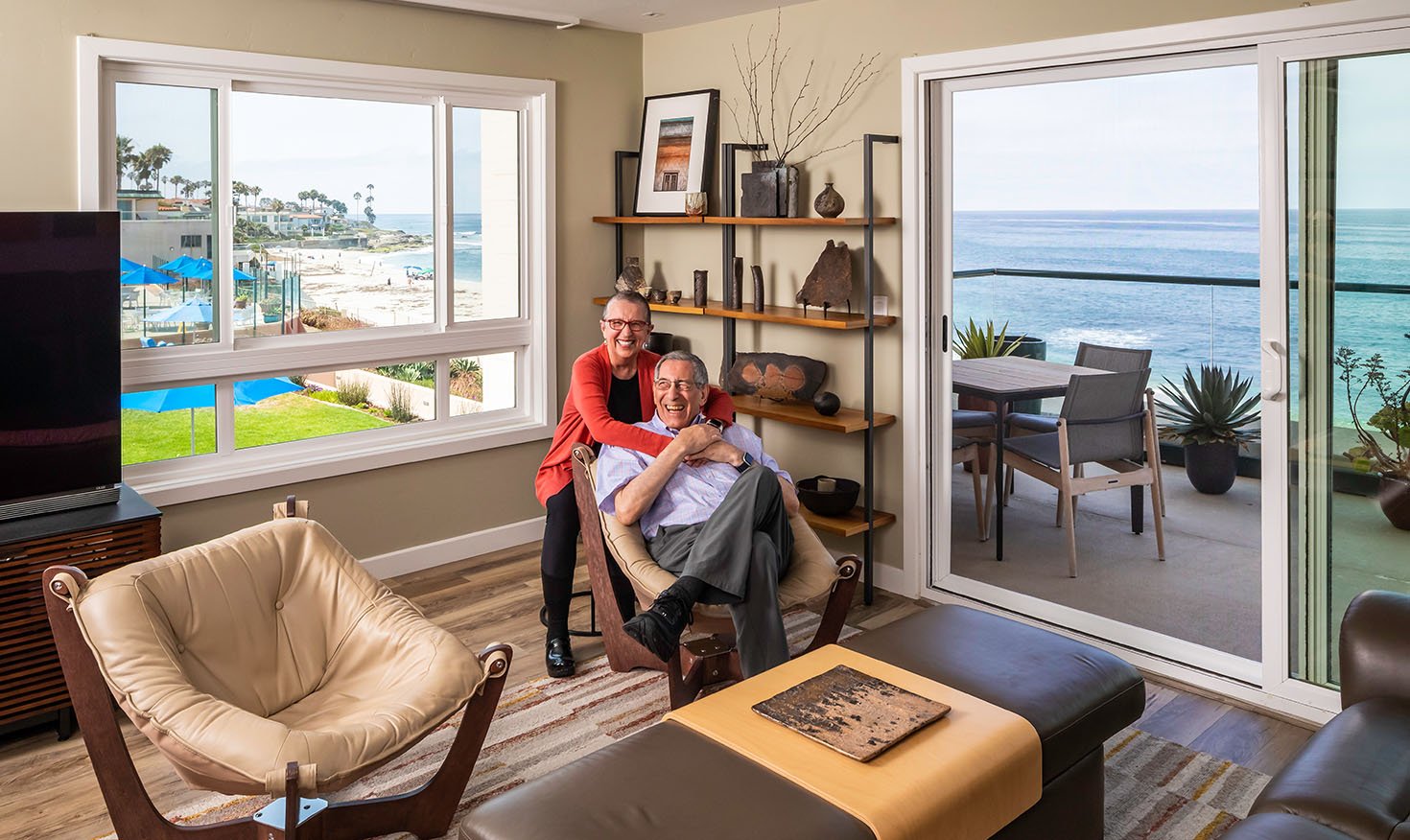 Senior woman with her arms around a senior man sitting inside their apartment with water views