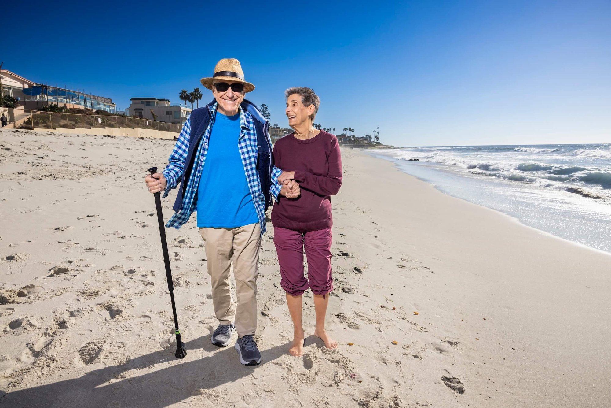 Senior couple walking on a sandy beach with ocean waves and clear sky.