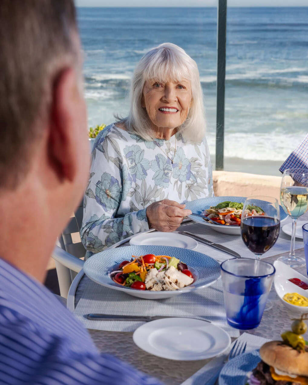 Senior woman dining outdoors by the ocean, with plates of food and drinks on the table