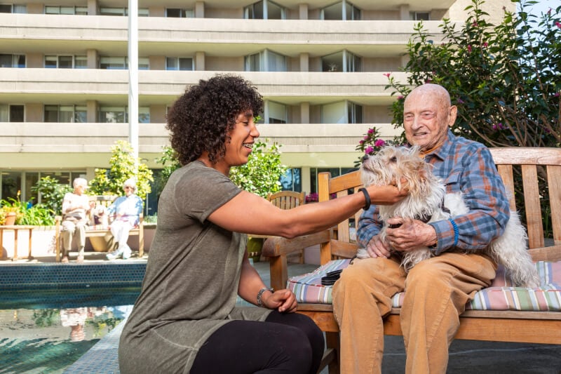 Woman petting a dog that's sitting on a senior man's lap on a bench outside Piedmont Gardens