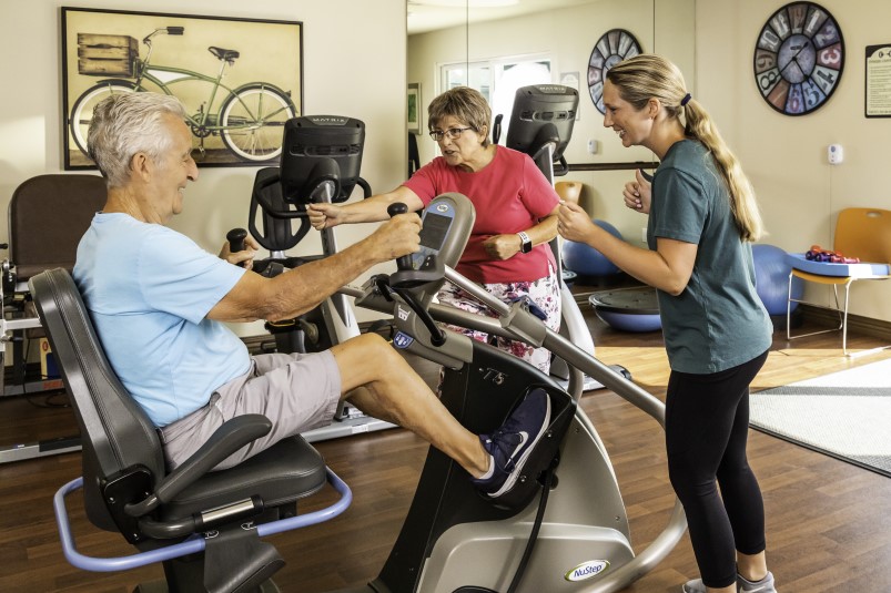 Senior man on recumbent bike while a senior woman and female personal trainer look on in the fitness center at Redwood Terrace