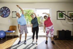 Senior man, senior woman and a female personal trainer do a side stretch in the fitness center at Redwood Terrace