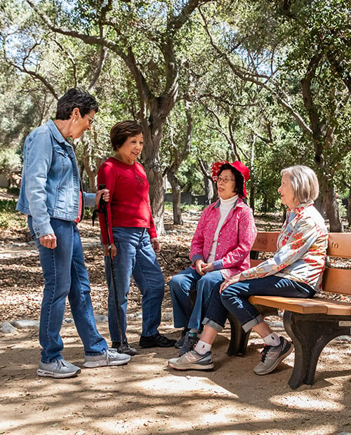 Two senior women standing and talking to two senior women sitting on a bench off a trail at Royal Oaks