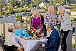 Group of seniors with wine glasses sitting at a table and standing on a balcony at Piedmont Gardens