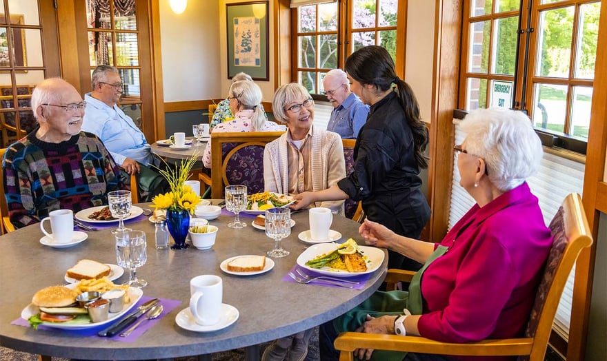 Dining room employee presenting plate of food to a senior woman sitting with two others in the dining room at The Terraces at Summitview