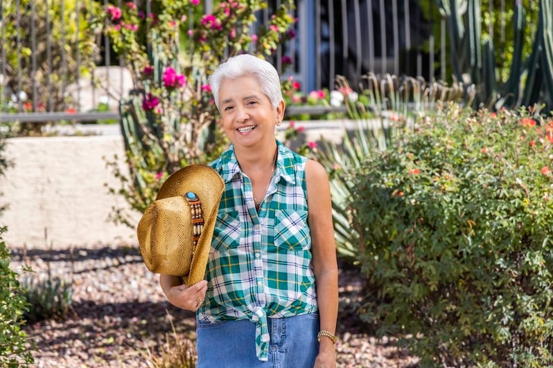 A smiling woman holding a cowboy hat in a garden with pink flowers and greenery.