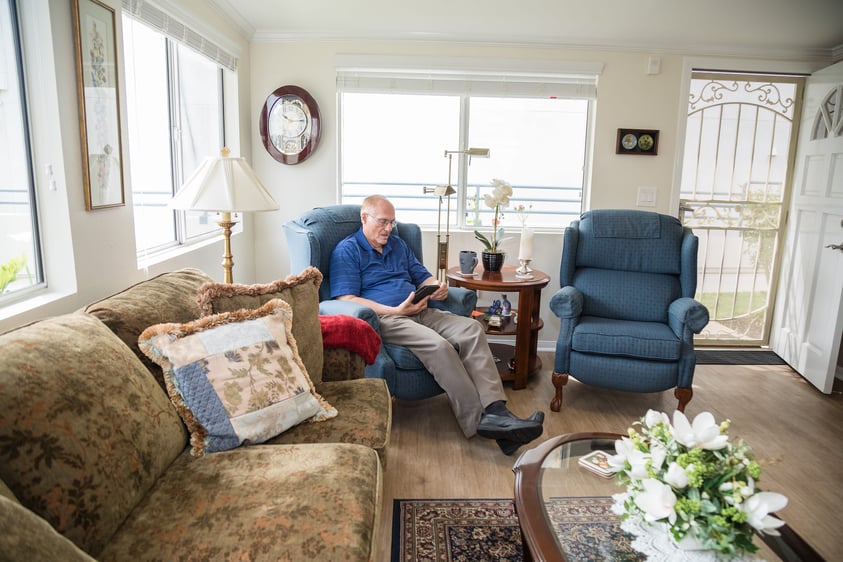 Senior man sitting in a blue chair reading in his home at Redwood Terrace