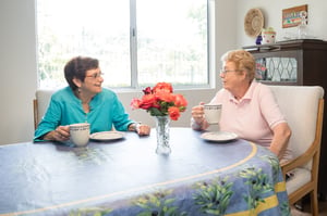 Two senior women drinking from mugs at the kitchen table of a home at Redwood Terrace