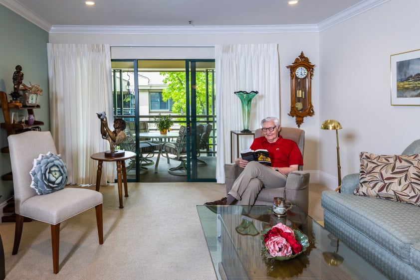 Senior man sitting in an armchair and reading a book in his apartment at The Terraces of Los Gatos