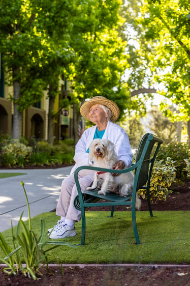 Senior woman wearing a sun hat while sitting on an outdoor bench and holding a small dog