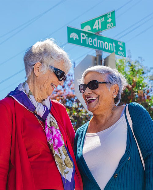 Two smiling senior women wearing sunglasses and standing at the corner of 41st Street and Piedmont Avenue