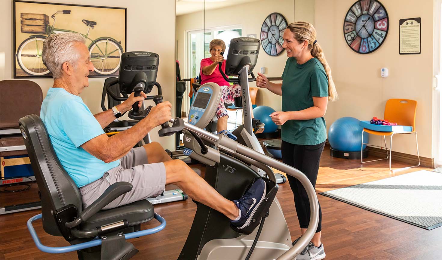 Senior man and senior woman using exercise machines in the fitness center at Redwood Terrace as a personal trainer looks on