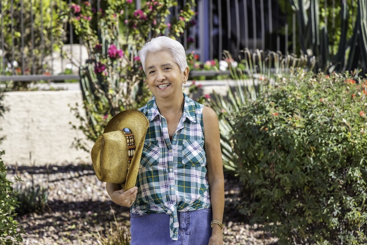 A smiling woman holding a cowboy hat in a garden with pink flowers and greenery.