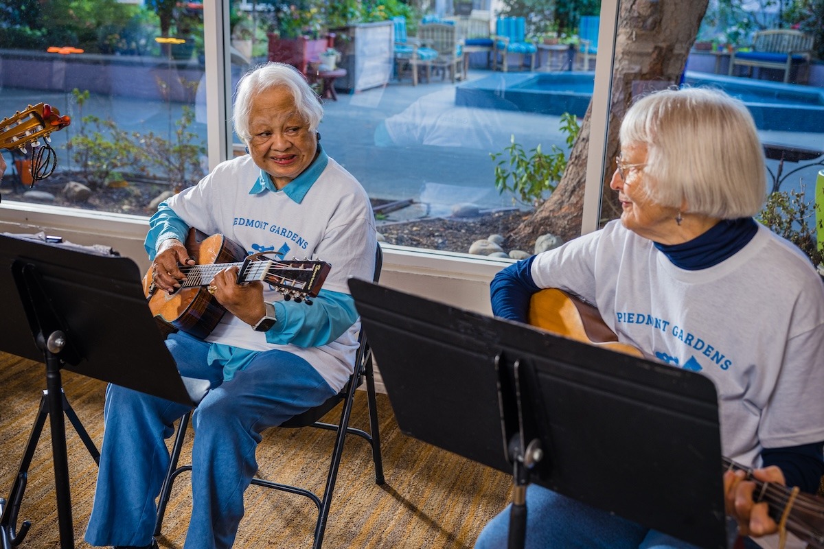 Two senior women playing guitars indoors, wearing matching 