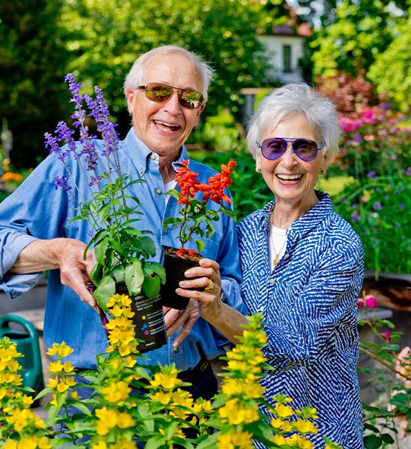 Smiling seniors, Mike and Linda, in a bright flower garden holding plants.