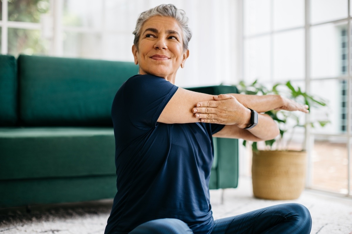 Older adult stretching indoors with a green sofa and potted plant in the background.